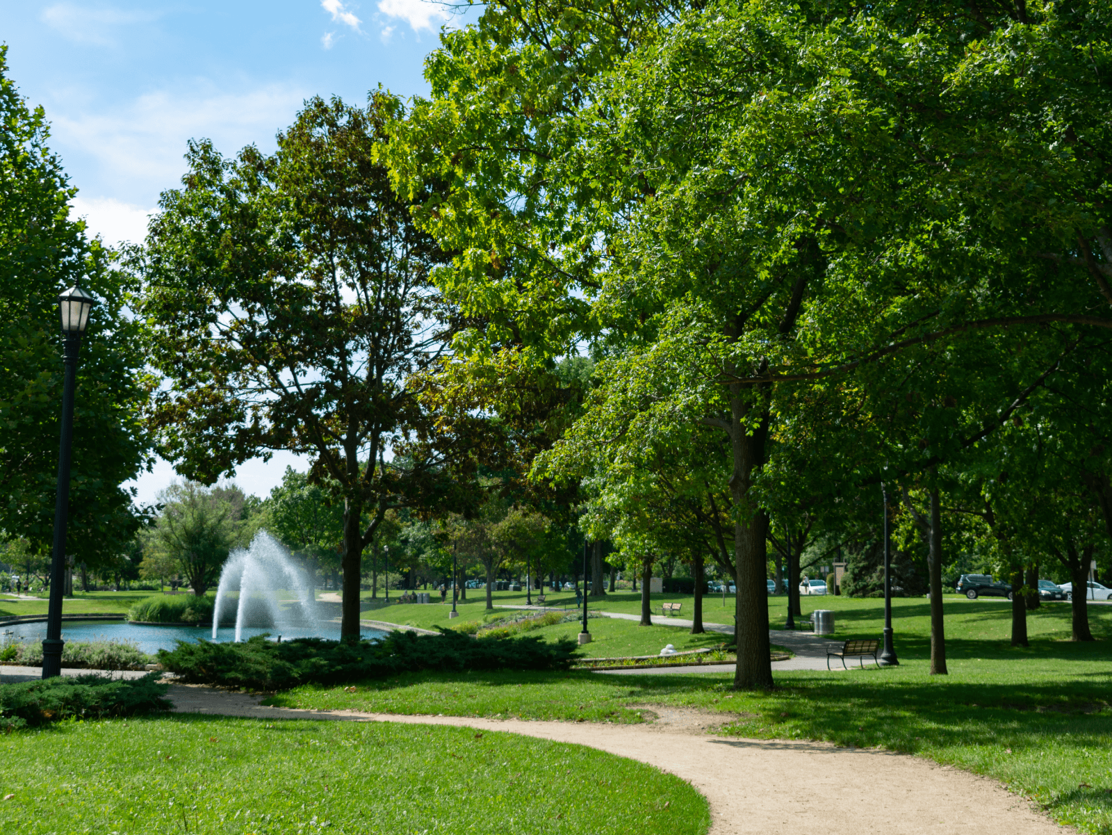 A photo of Evanston, Illinois that is taken in a park. There is a pond with a fountain in the background, with a path leading to it in the foreground. The trees are lush and green.