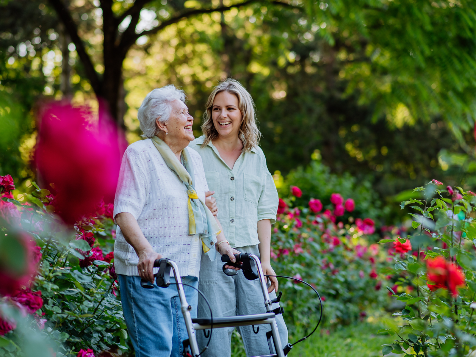 A woman and her elderly mother are walking through a garden with pink and red flowers. The mother is using a walker.