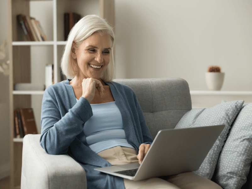 An older white woman smiling while looking at her laptop and sitting on the couch