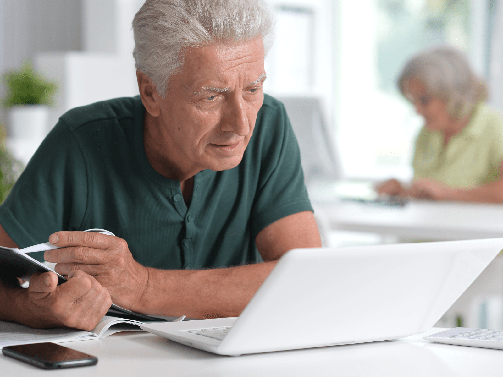 Older white man wearing a green shirt sits at a table with his laptop open, looking confused