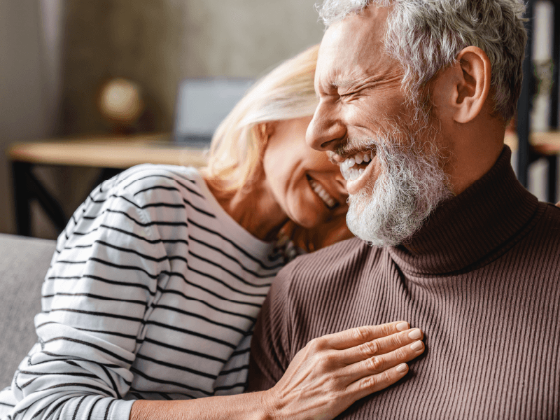 An older man and woman lean into each other while smiling and laughing. The woman has her hand on his chest.